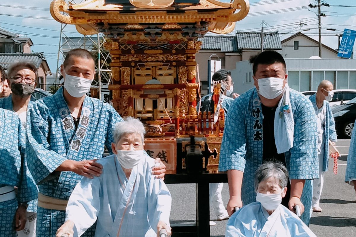 地域のお祭りで神輿の前での記念写真、幸田健太と若い男性、年老いた女性2人が写っている